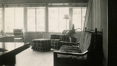 Scene from the video showing the interior of the house, with chairs against the wall and windows in the background.