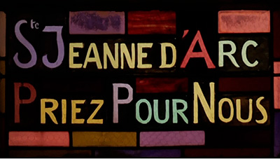 Scene from the video showing the detail of a stained-glass window where we can read «St Jeanne d'Arc, pray for us».