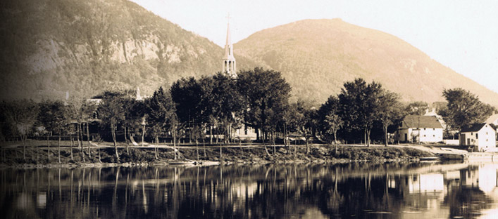 View of Saint-Hilaire from accross the Richelieu river with the church, trees and the mountain in the background.